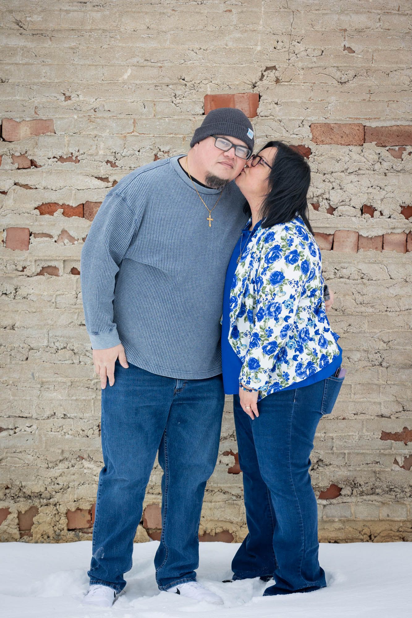 Affectionate couple poses together in front of a rustic brick wall in winter snow.