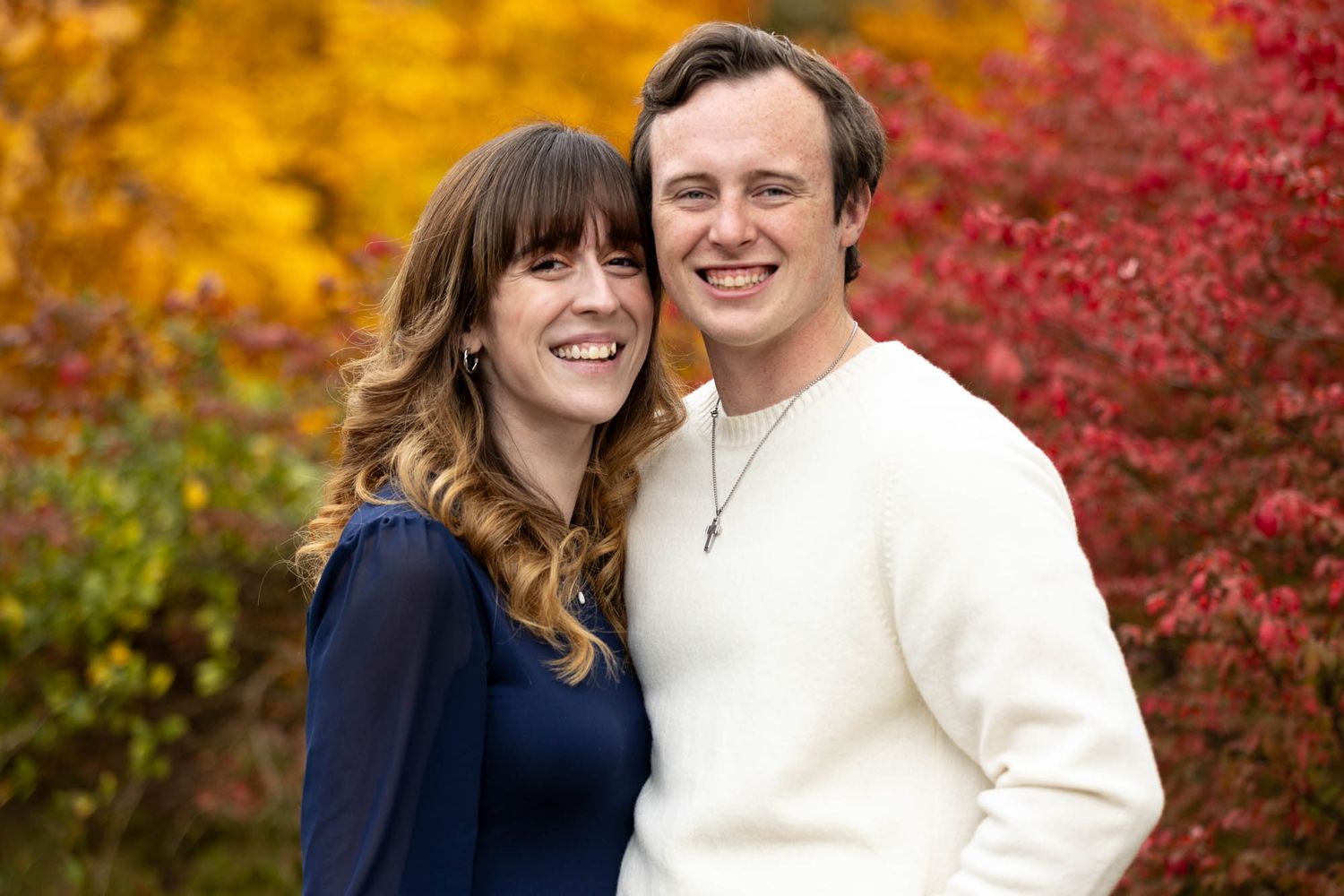 Smiling couple poses together in front of vibrant autumn foliage during fall engagement photo session.