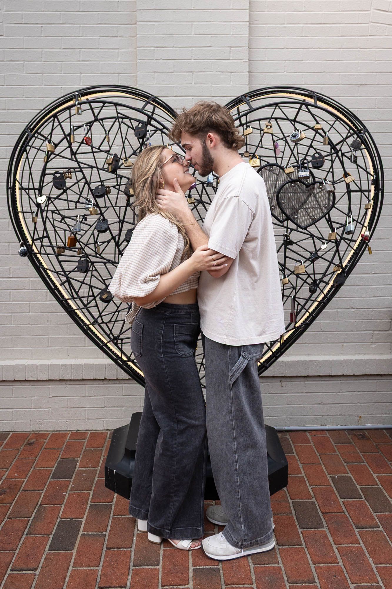 A romantic couple embraces in front of a large decorative heart sculpture on a brick patio.