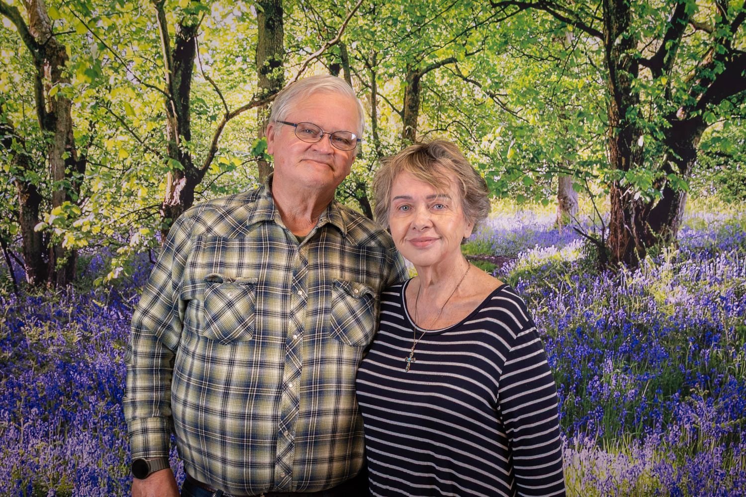 Senior couple smiling together in front of scenic bluebell woodland forest backdrop.