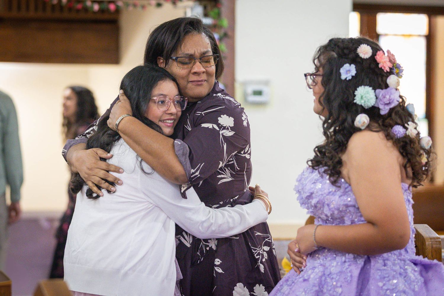 Woman in purple dress embraces two girls in church during a quinceañera celebration.