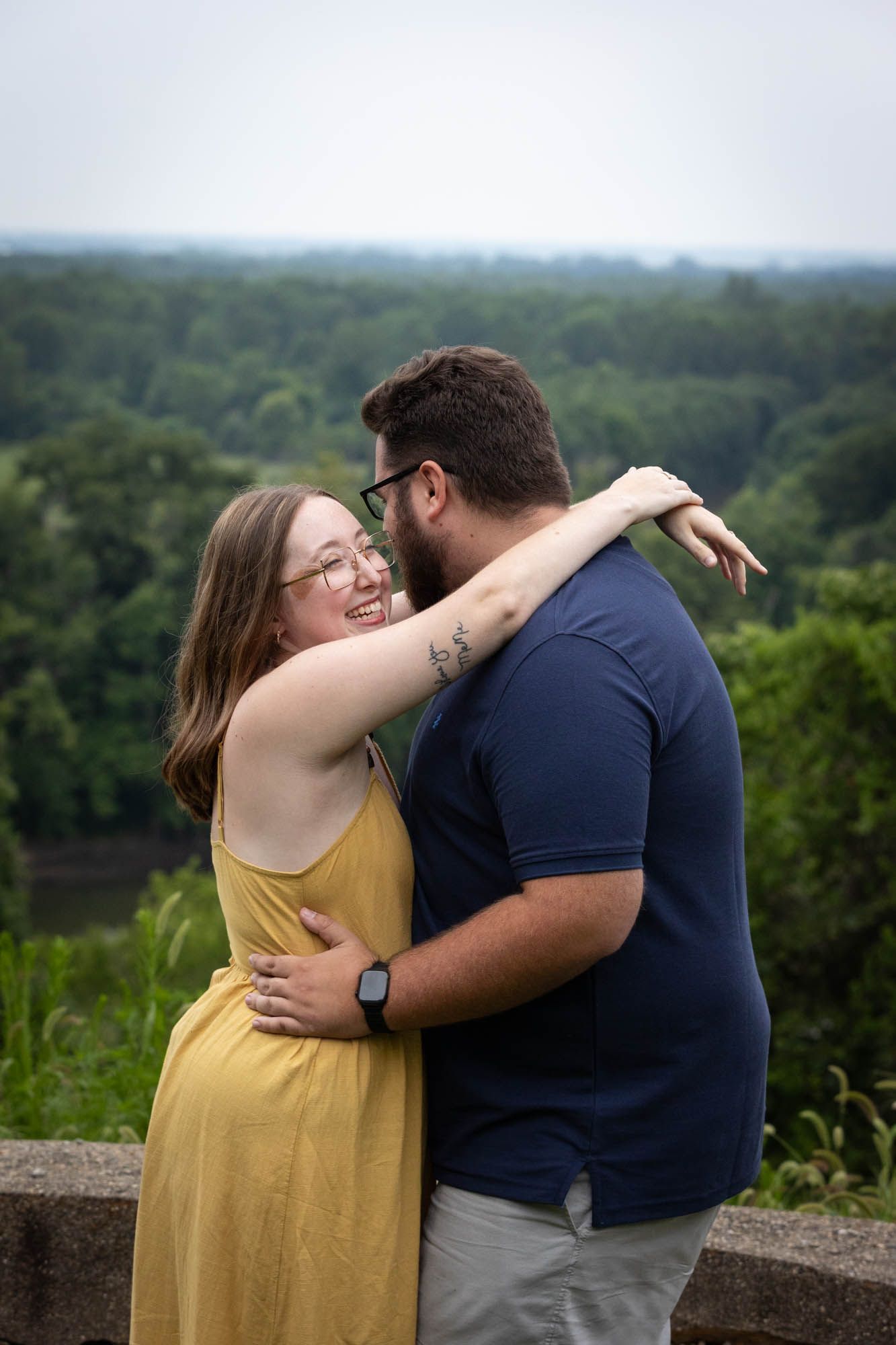 A couple embraces and smiles outdoors, with a lush green forest landscape in the background.