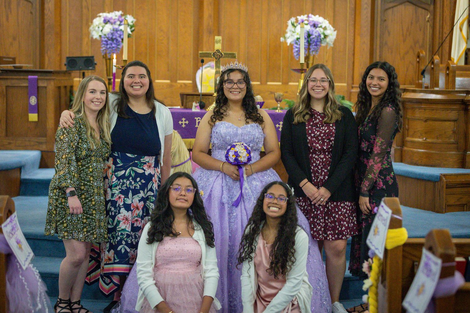 Quinceañera honoree in lavender gown poses with her chambelanes and damas inside a decorated church.