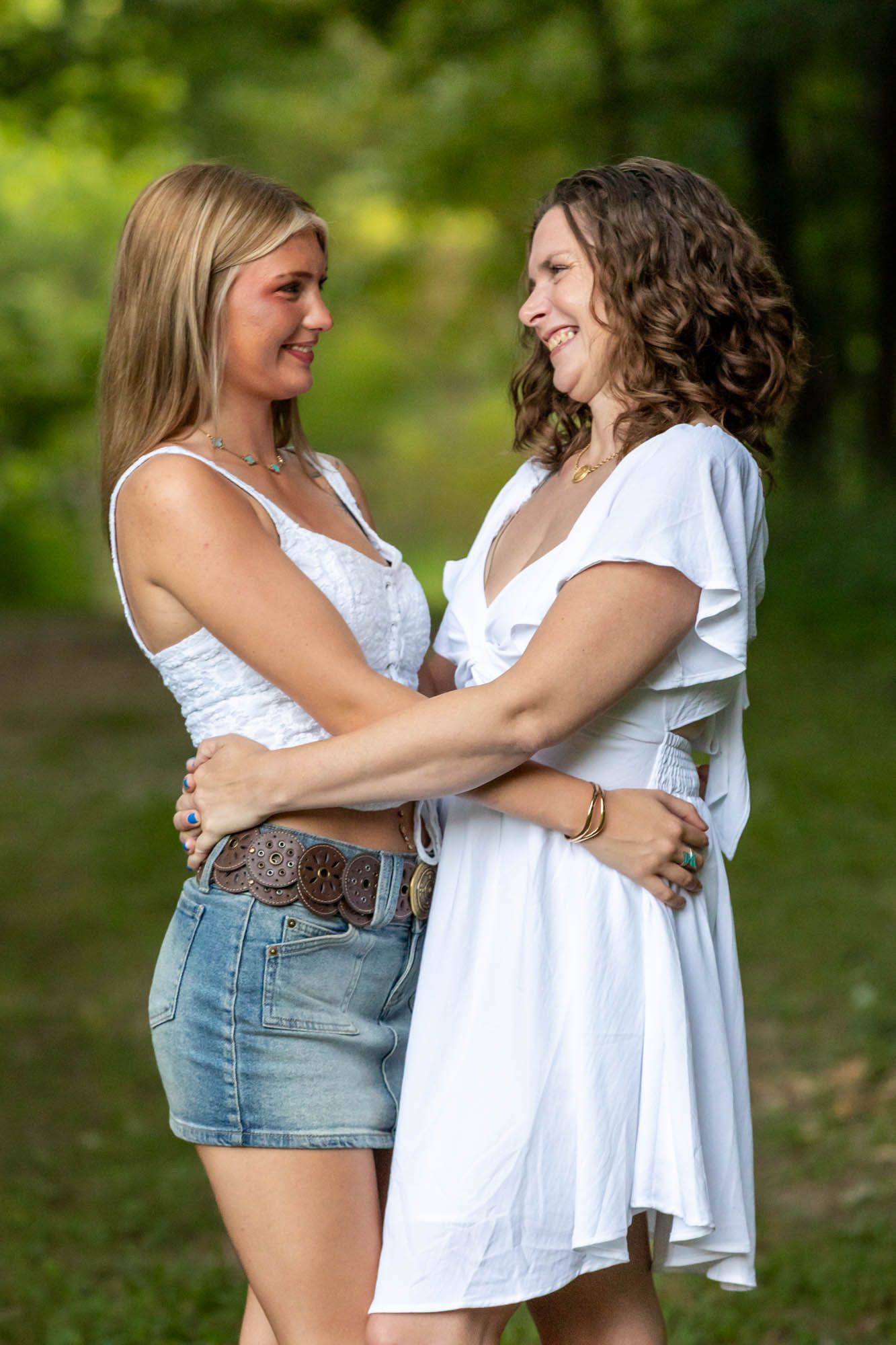Two smiling women in white tops embrace outdoors in a lush green park setting.
