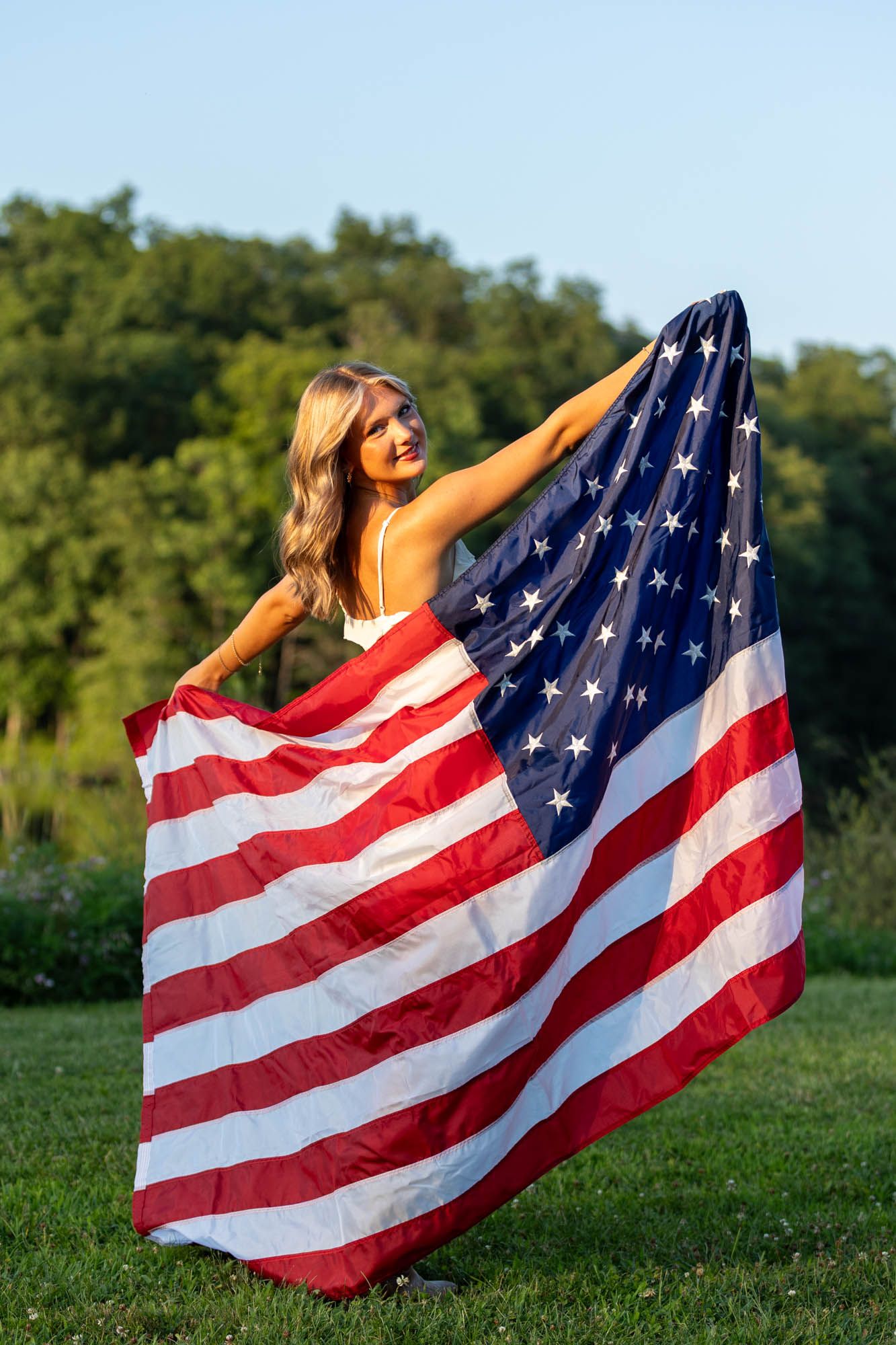 Young blonde woman holding a large American flag outdoors on green grass with trees in background.