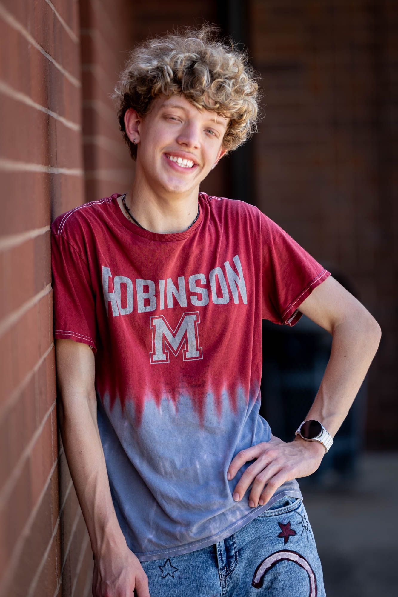 Smiling teen with curly blond hair wearing a Robinson M graphic tee leans against a brick wall outdoors.