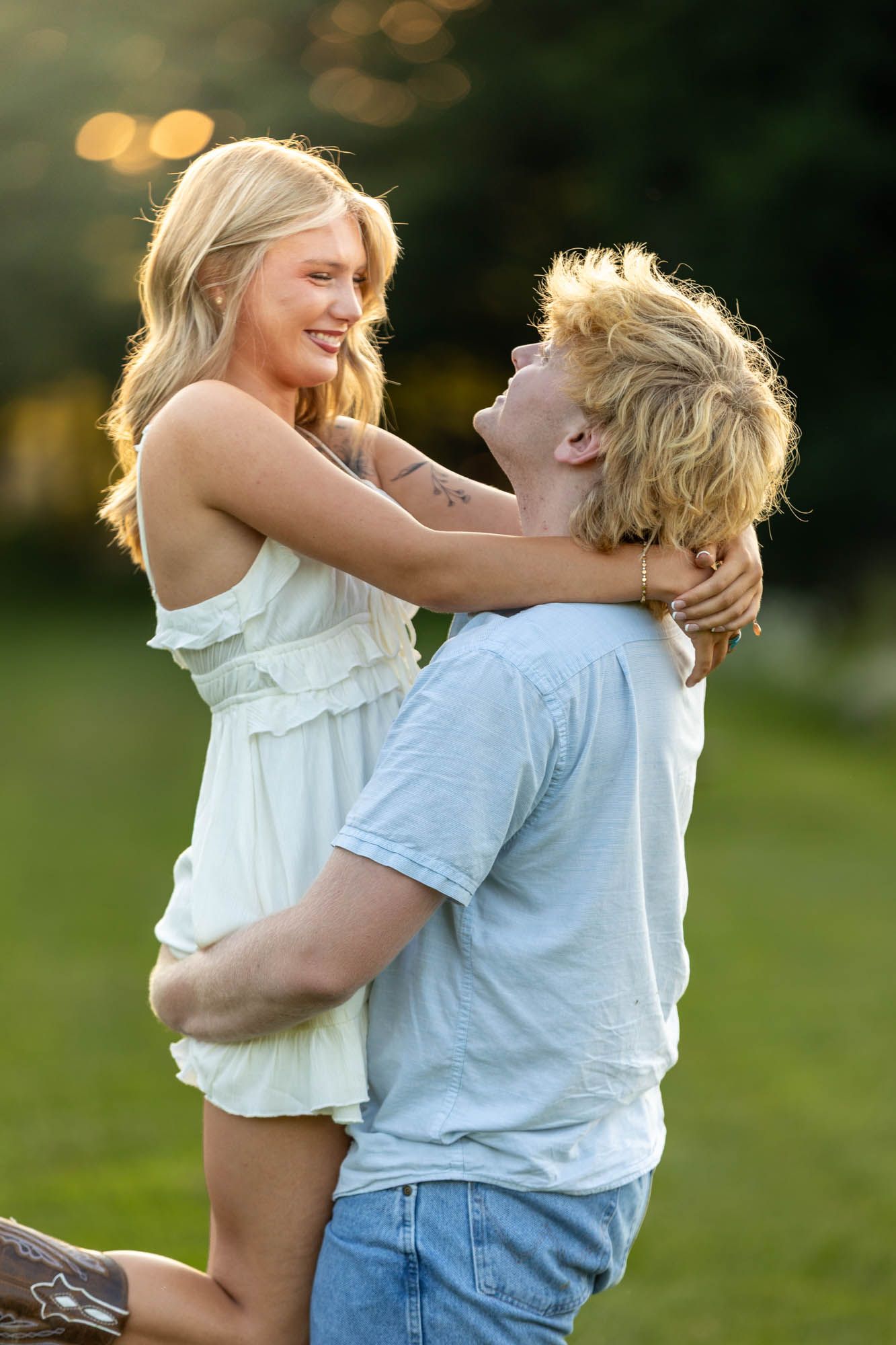 Young couple embracing outdoors at golden hour, woman in white dress lifted by man in blue shirt on green lawn.