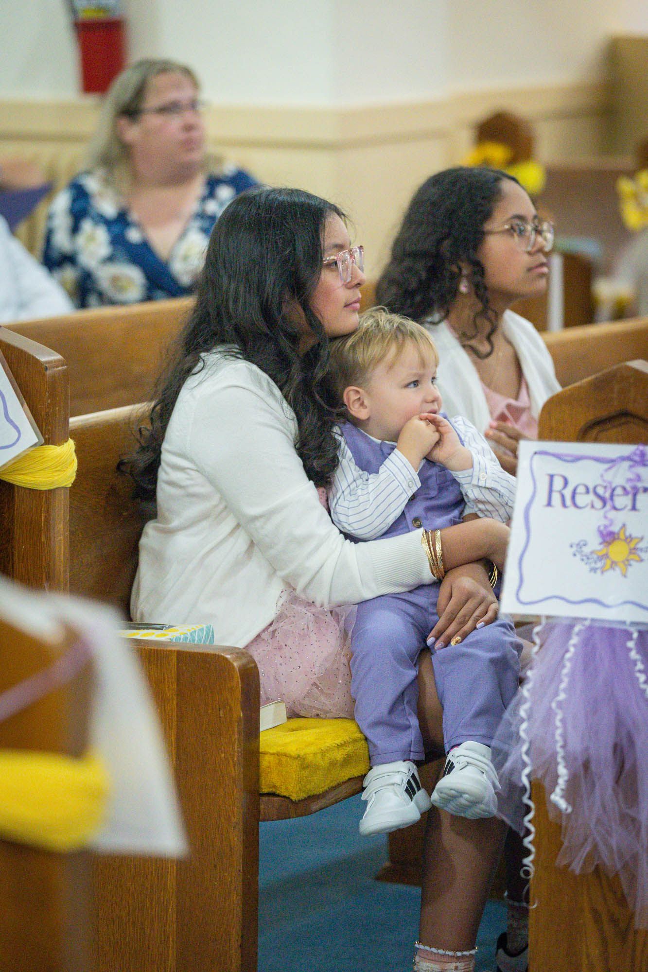 Family members seated in wooden church pews watching a quinceañera ceremony with a reserved sign visible.