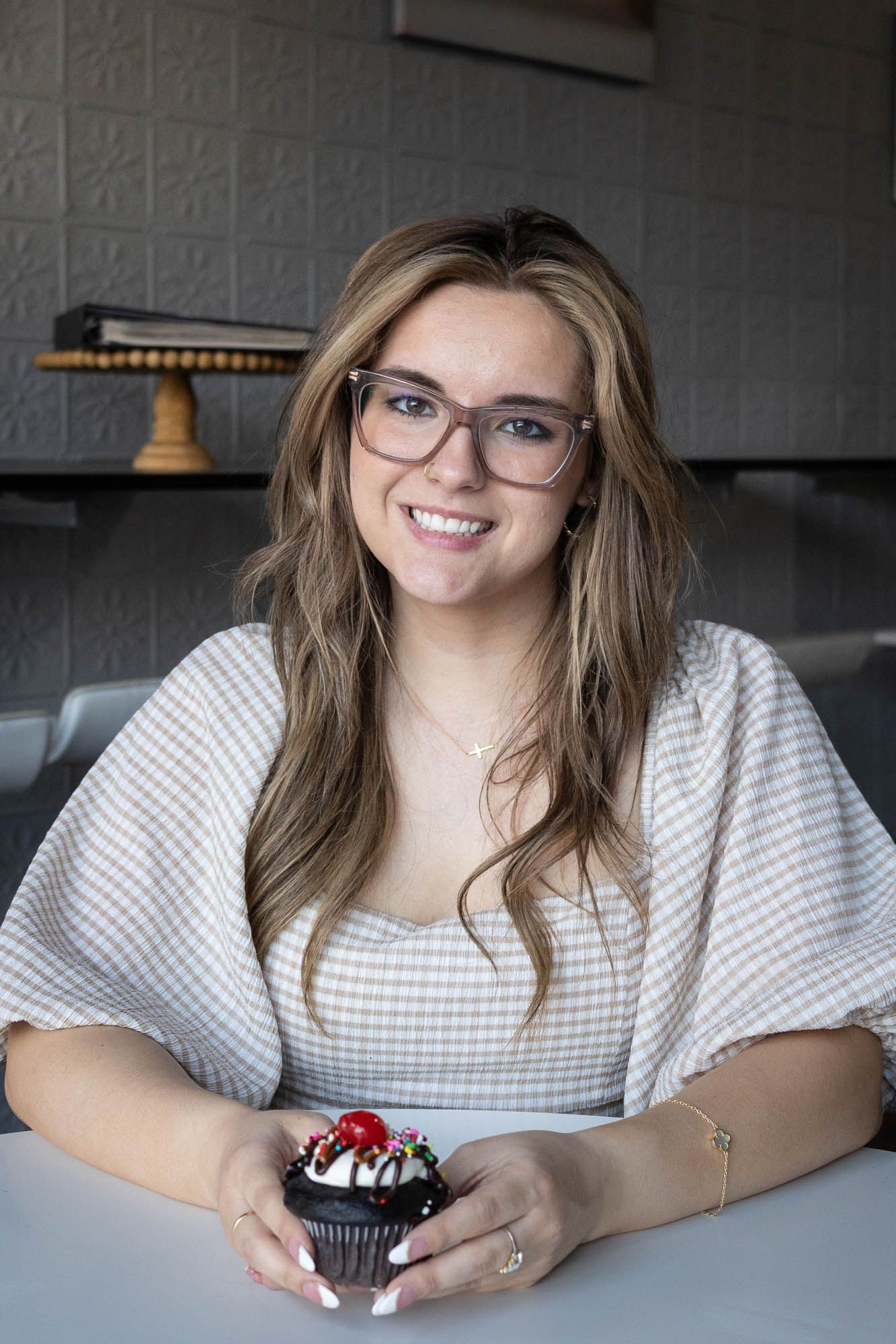 Smiling young woman with glasses holding a chocolate cupcake with cherry topping at a cafe table.