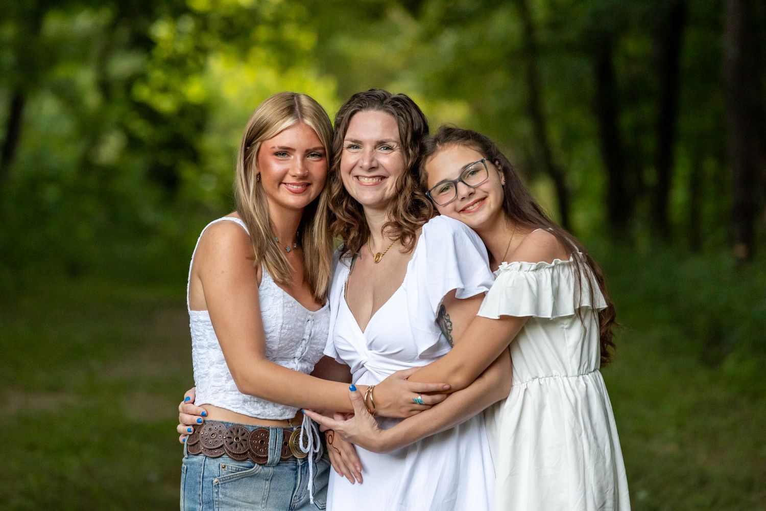 Three smiling women in white outfits embrace outdoors in a lush green forest setting.