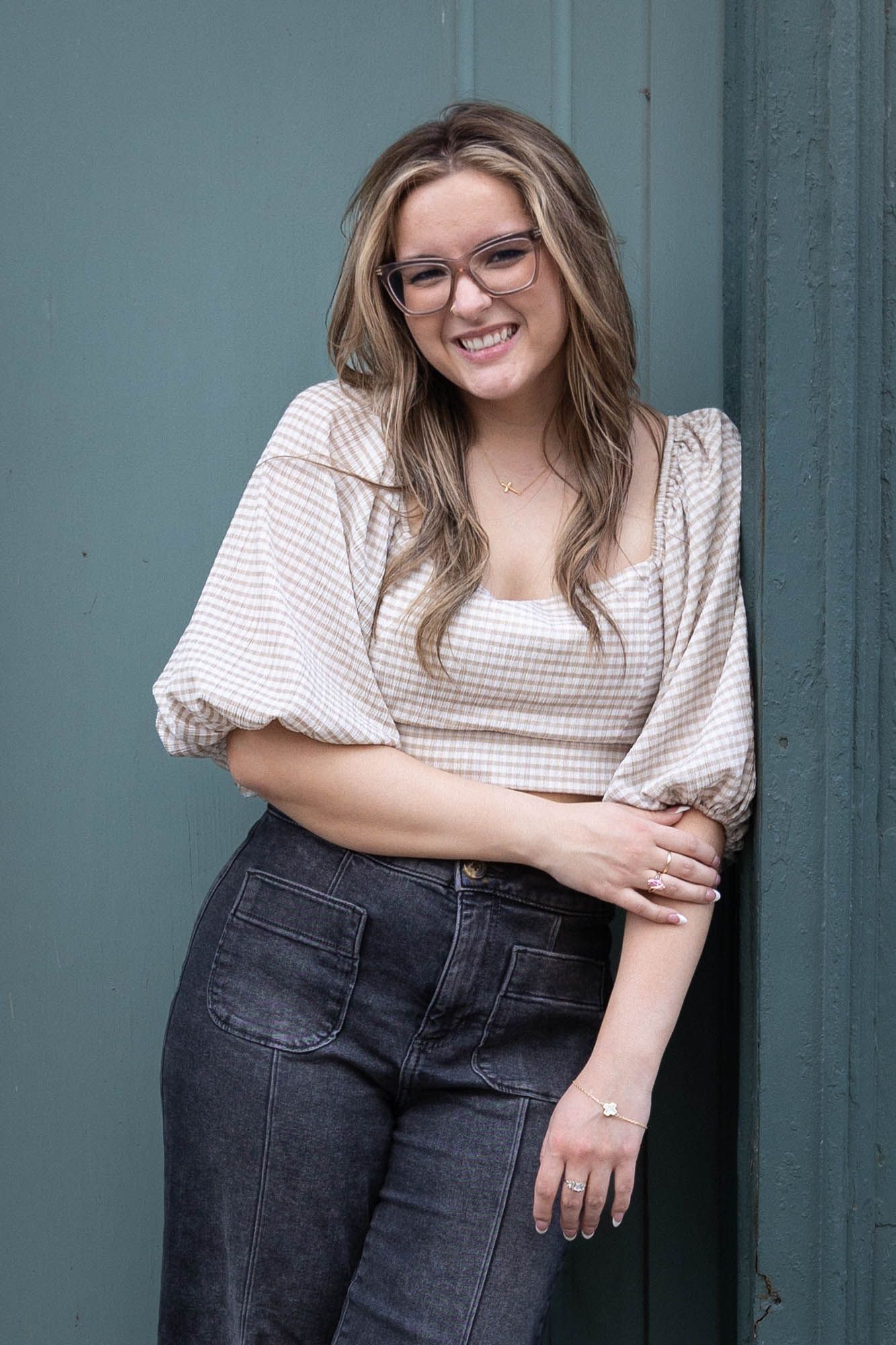 Happy young woman with glasses smiling and leaning against a teal door in a checkered crop top.