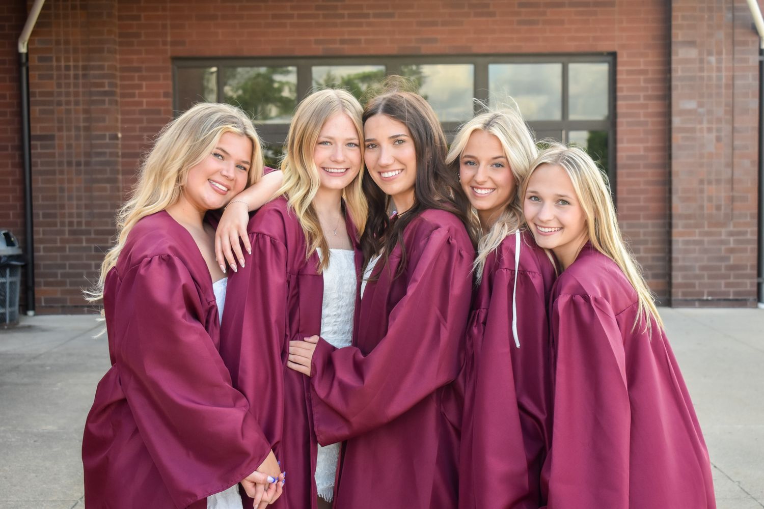Five young women in maroon graduation gowns pose together smiling outside a brick building.