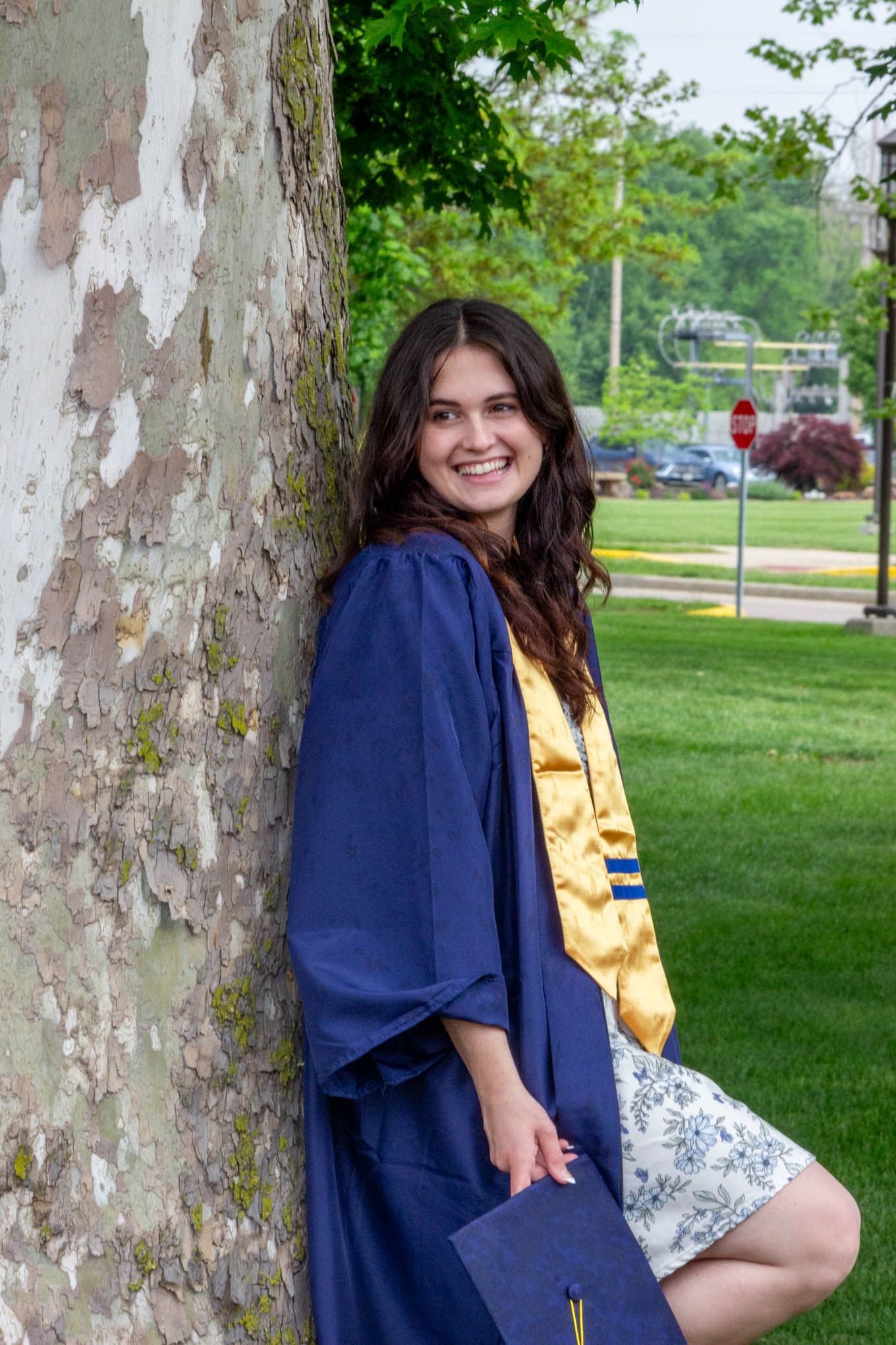 Smiling graduate in blue gown and gold stole leans against a tree holding her graduation cap.