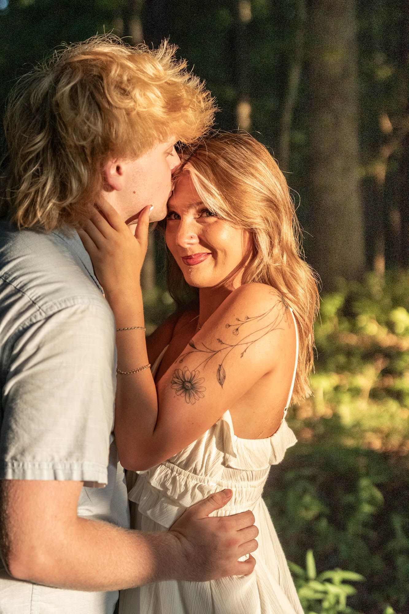 A couple shares a romantic moment outdoors, woman in white dress with floral tattoo smiles while embracing a blond man in golden light.
