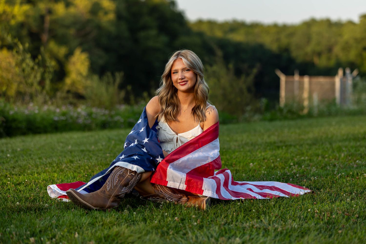 Young blonde woman wrapped in American flag sitting on green grass outdoors, smiling in golden hour light.