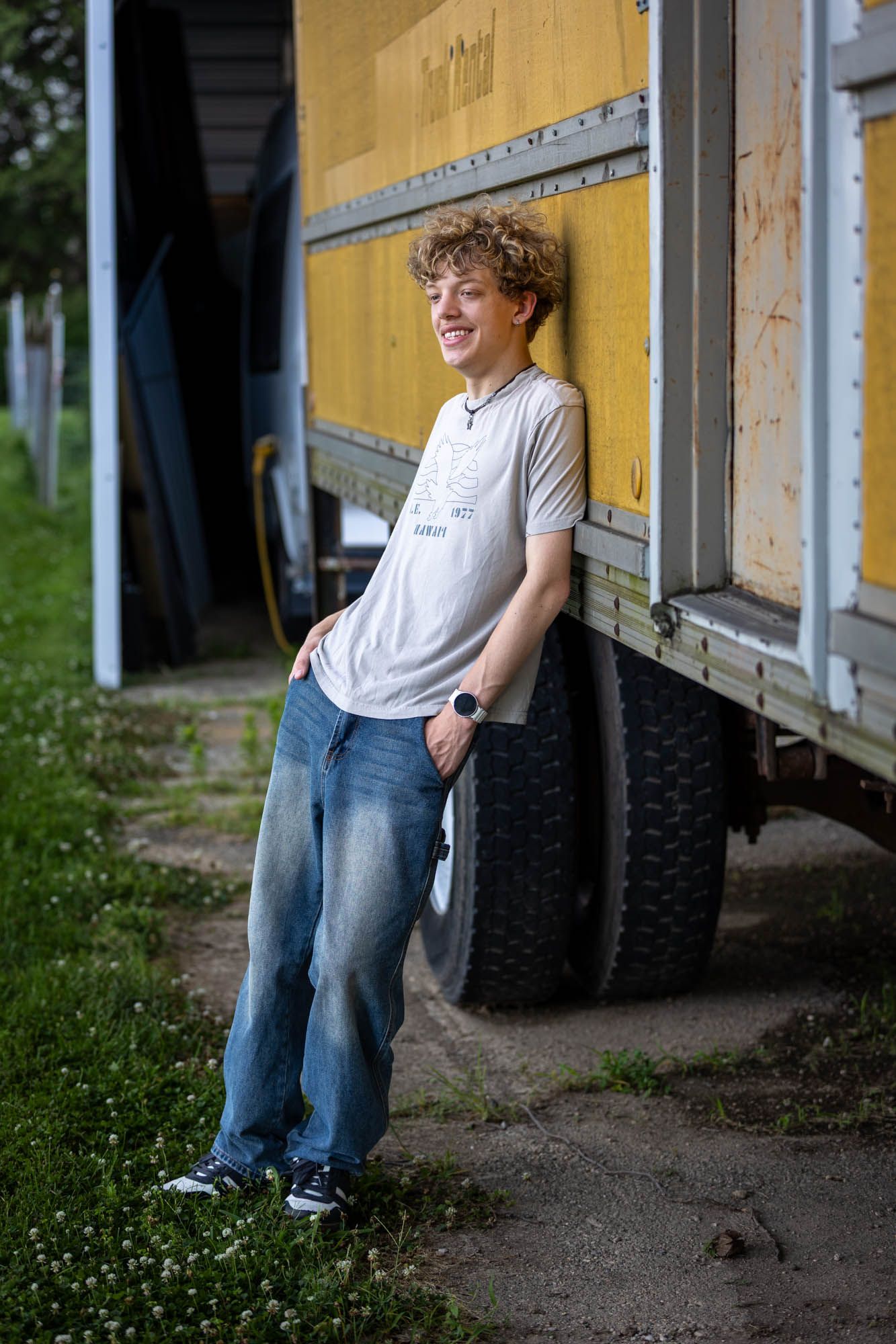Smiling teenage boy with curly hair leaning against a yellow truck, wearing jeans and a white t-shirt outdoors.