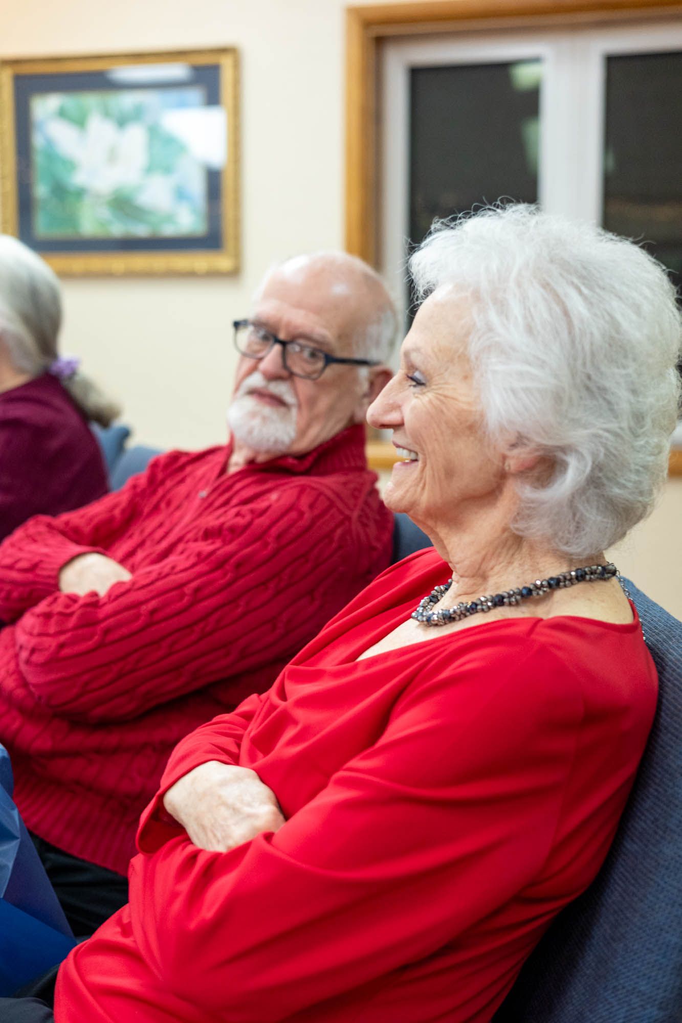 A smiling senior woman in red with white hair sits beside an older bearded man in a red sweater indoors.