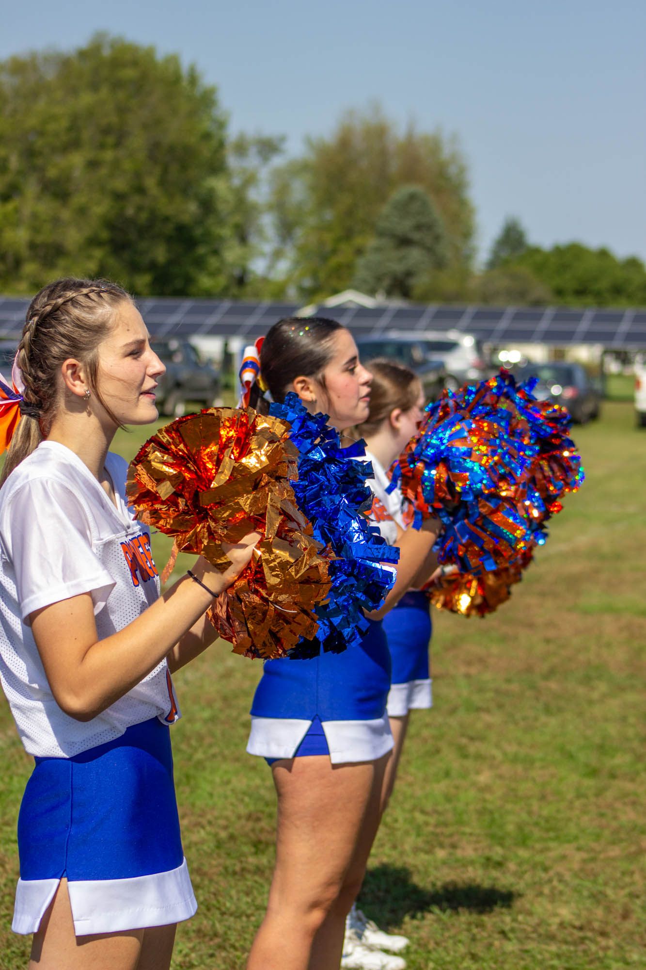 Cheerleaders in blue and white uniforms hold orange and blue pom-poms while cheering at an outdoor football game.
