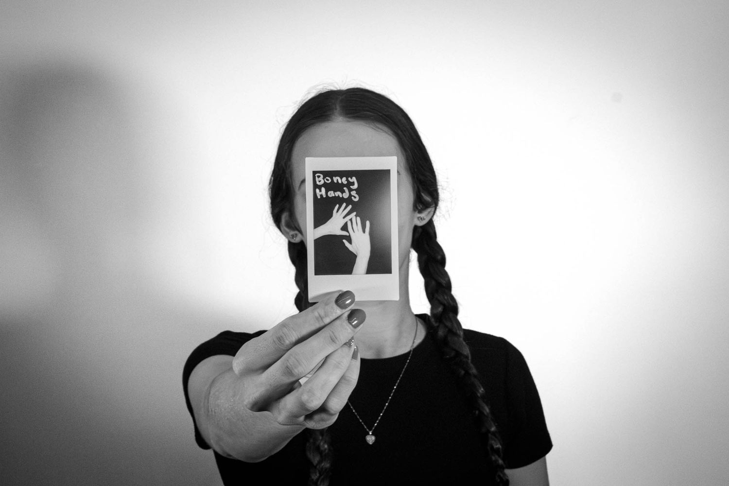 Black and white portrait of a girl with braids holding a Polaroid photo labeled Dummy Hands.