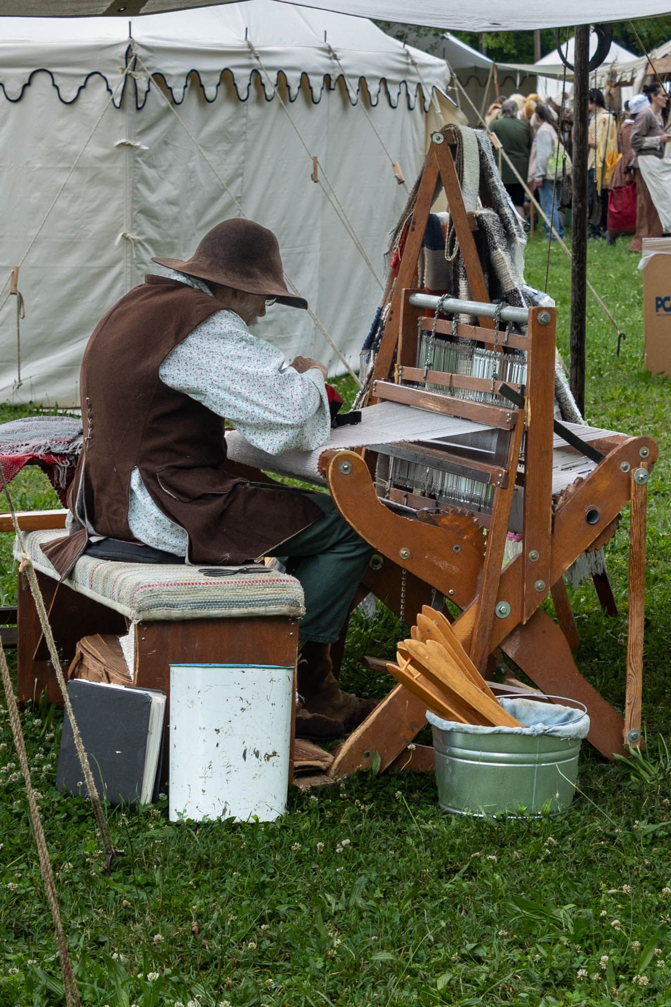 Historical reenactor in brown hat and period clothing operating a traditional wooden weaving loom outdoors.