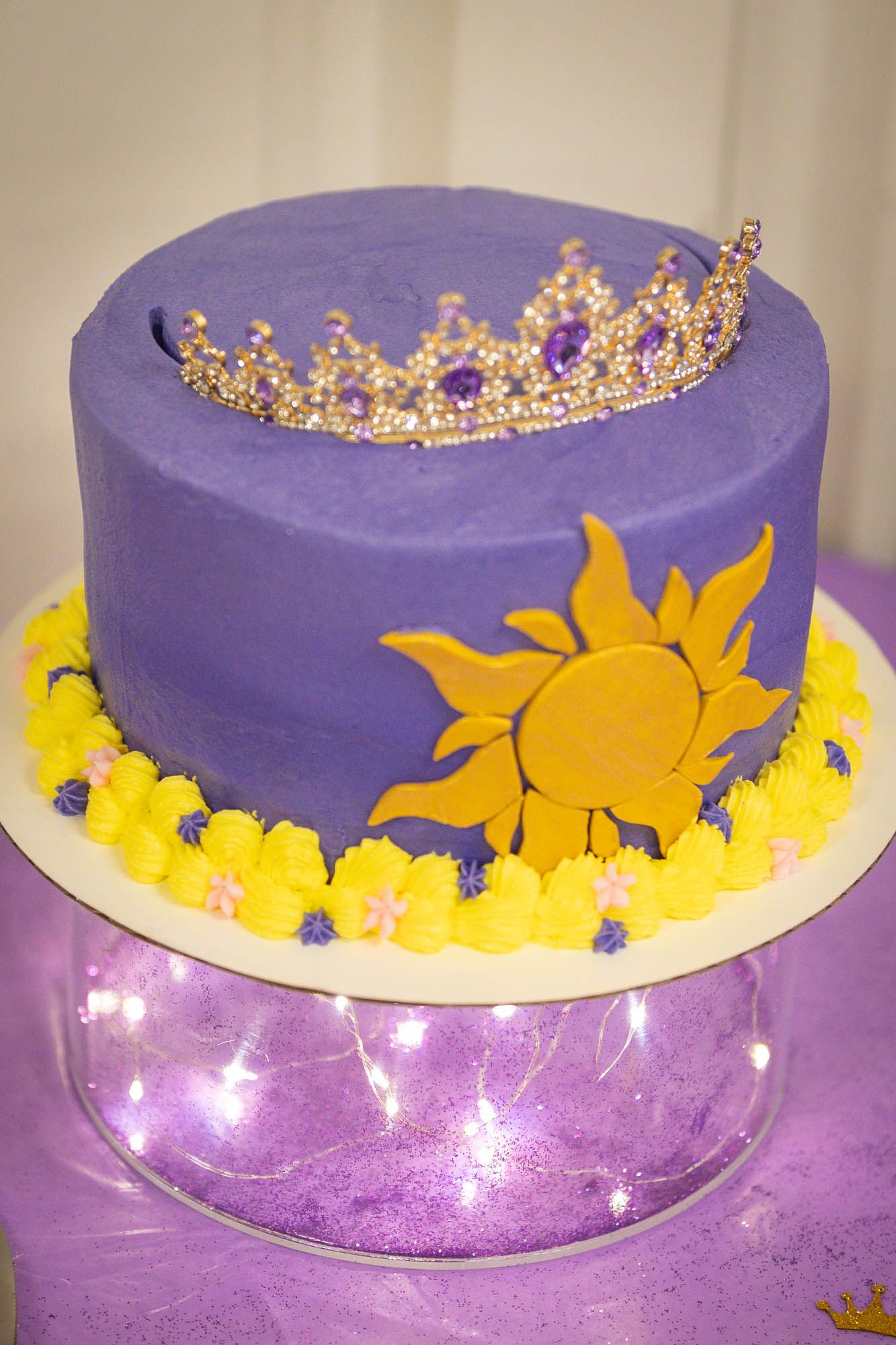Purple frosted quinceañera cake decorated with a gold sun, flowers, and a jeweled tiara on top.