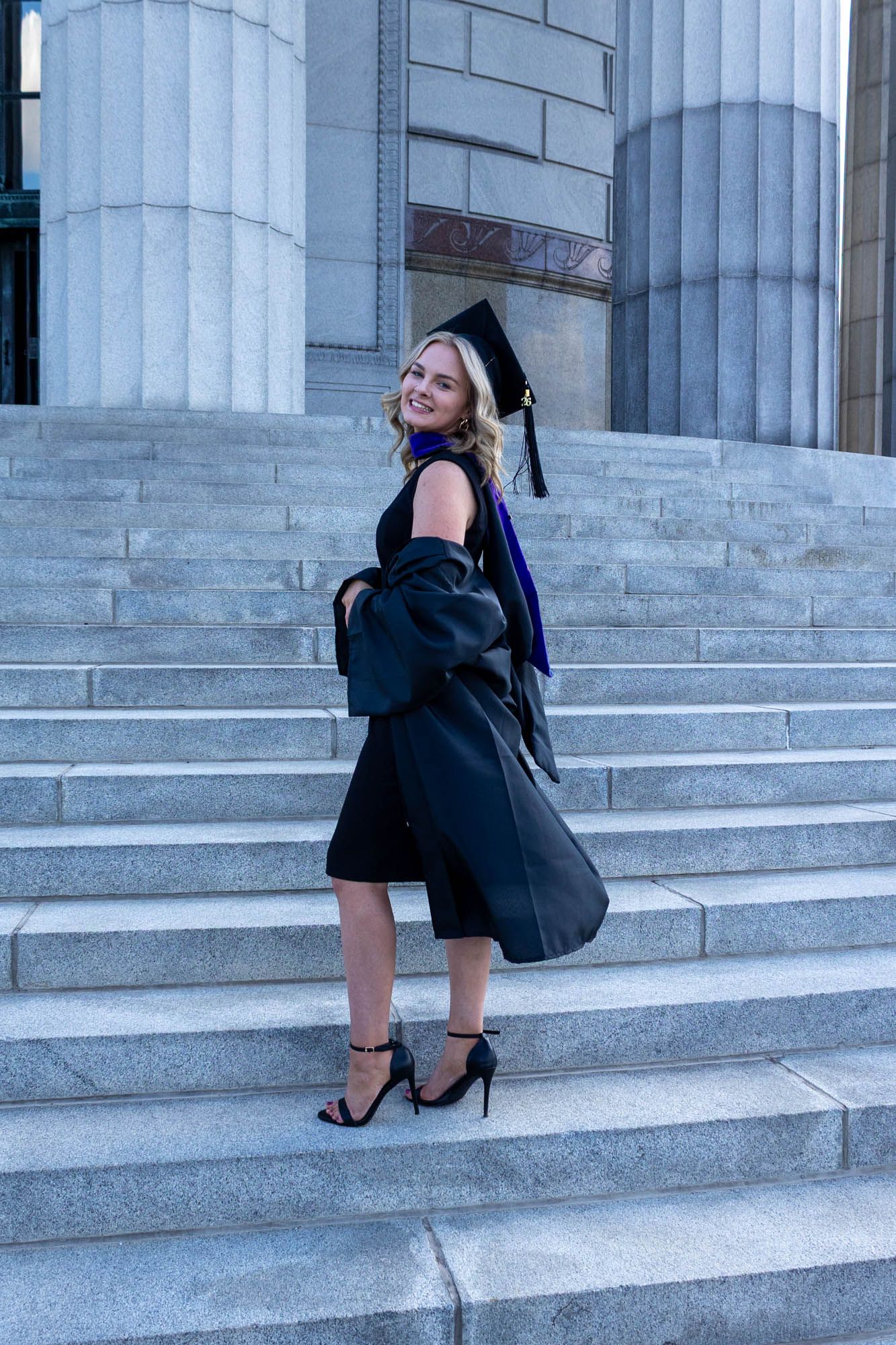 Blonde woman in black graduation cap and gown smiling on marble university steps in heels.