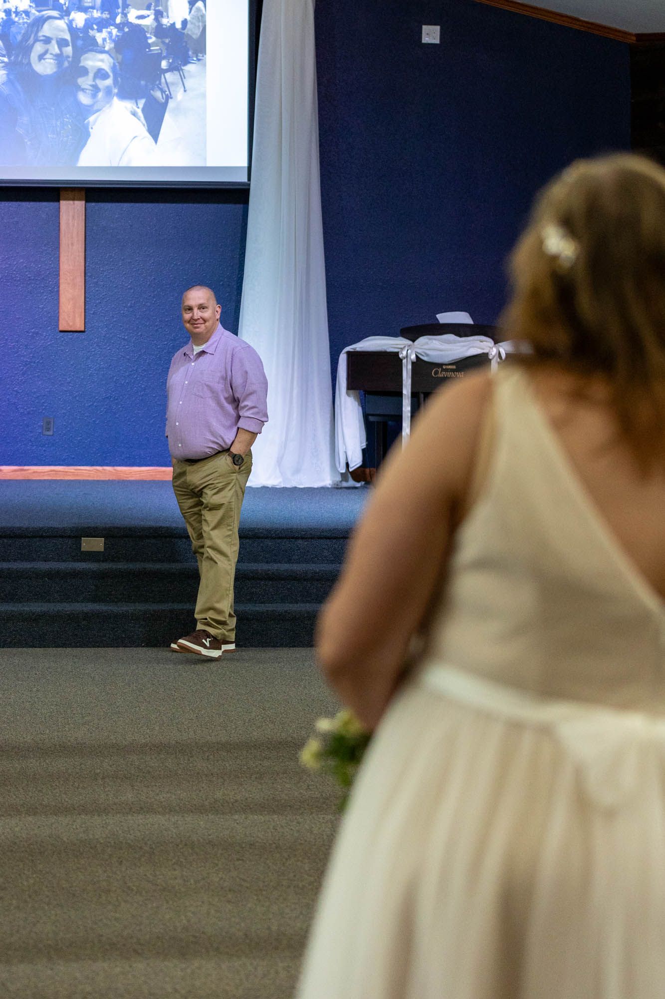 A man in a purple shirt smiles at a bride in a white dress during a wedding ceremony in a blue hall.