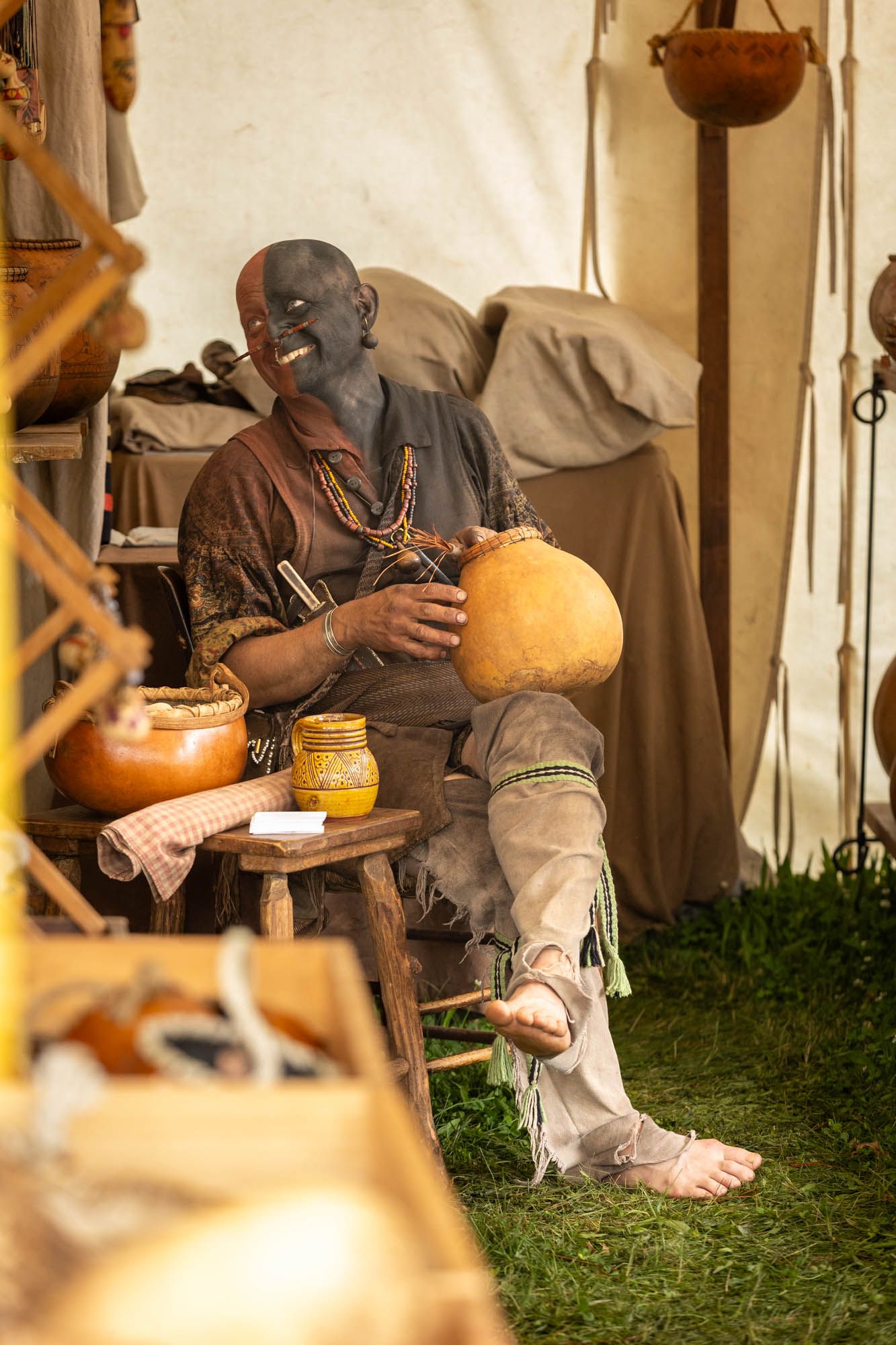 Medieval reenactor with face paint sits in tent holding gourd at historical festival.