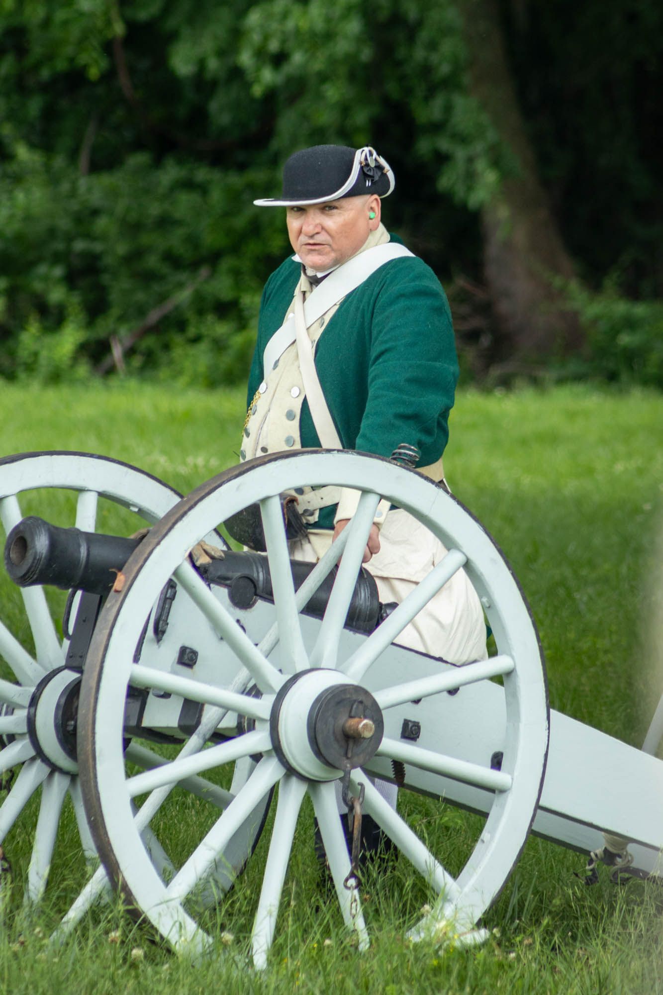 Revolutionary War reenactor in green uniform stands behind historic cannon on grass field.