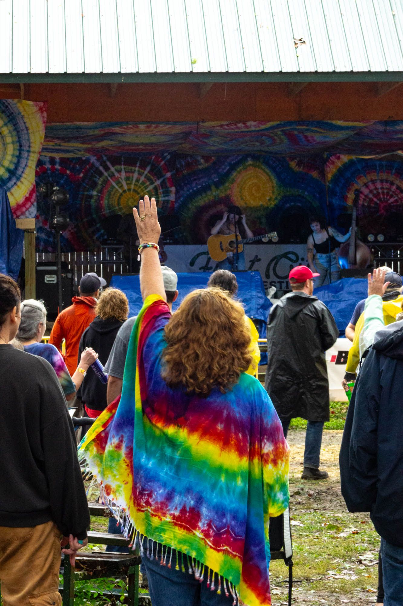 Woman in rainbow tie-dye shawl raises hand toward colorful outdoor concert stage.