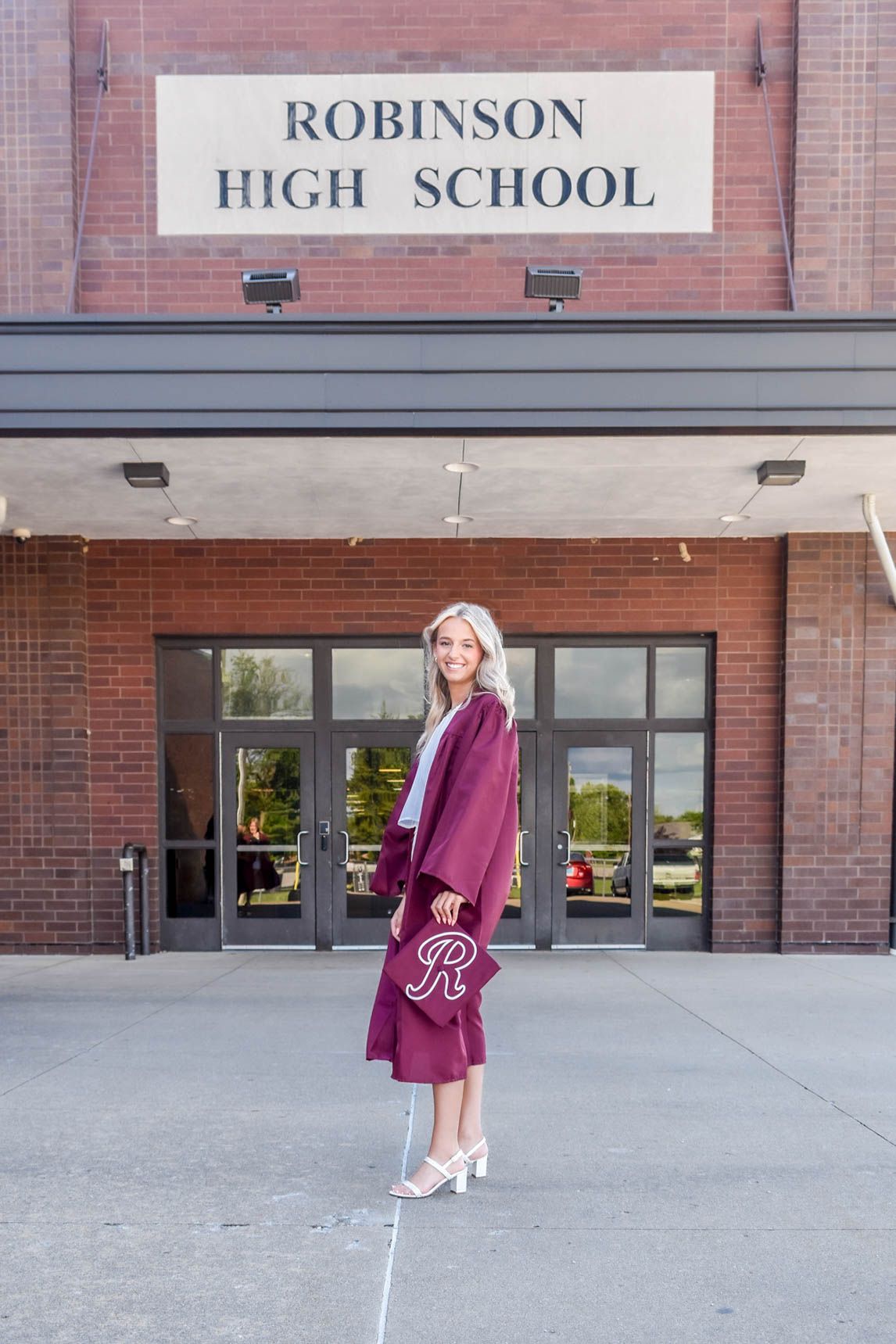A blonde female graduate in maroon cap and gown holds a decorated mortarboard outside Robinson High School.