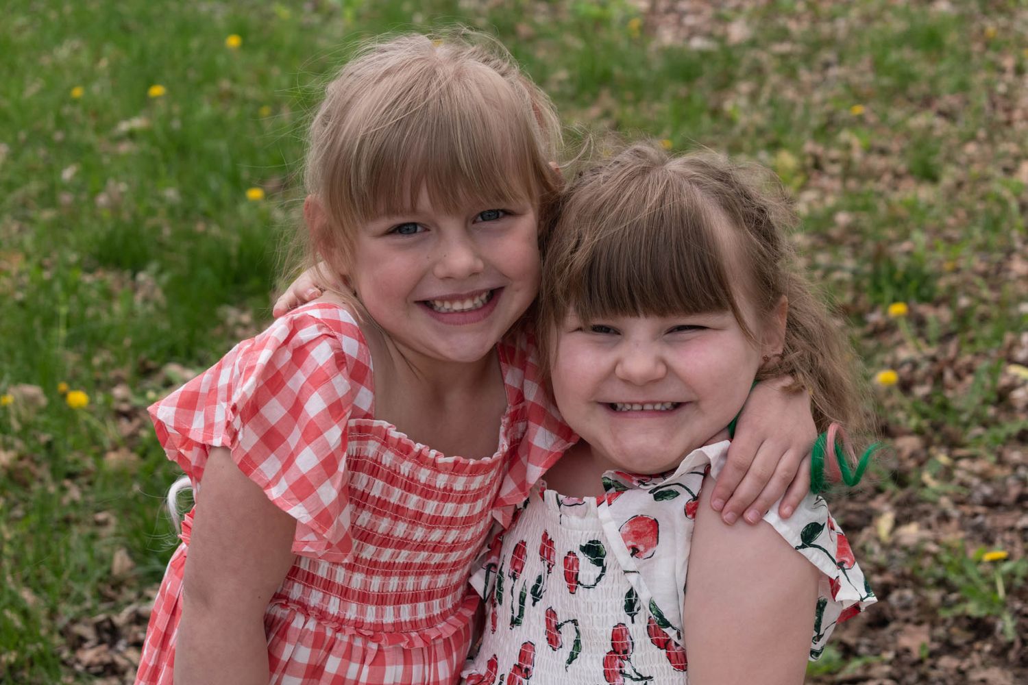 Two young girls in summer dresses share a warm hug while smiling outdoors on green grass.