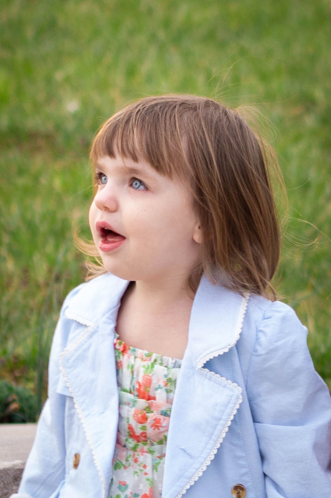 Cute toddler girl in light blue jacket and floral dress sitting outside with mouth open in surprise.
