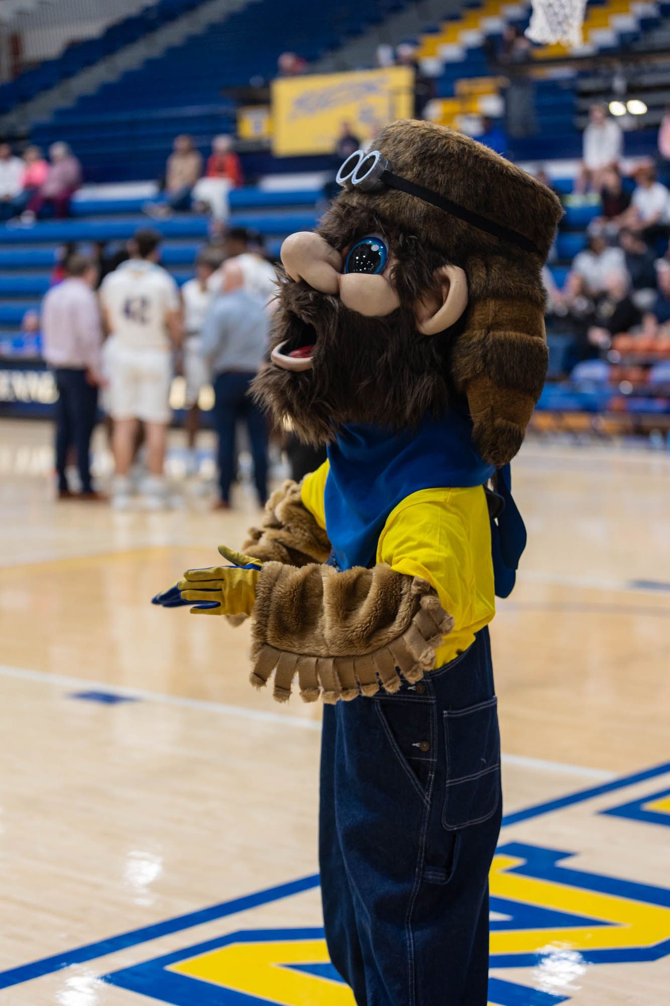 Vincennes University furry mascot in blue overalls and yellow shirt entertains crowd at basketball game.
