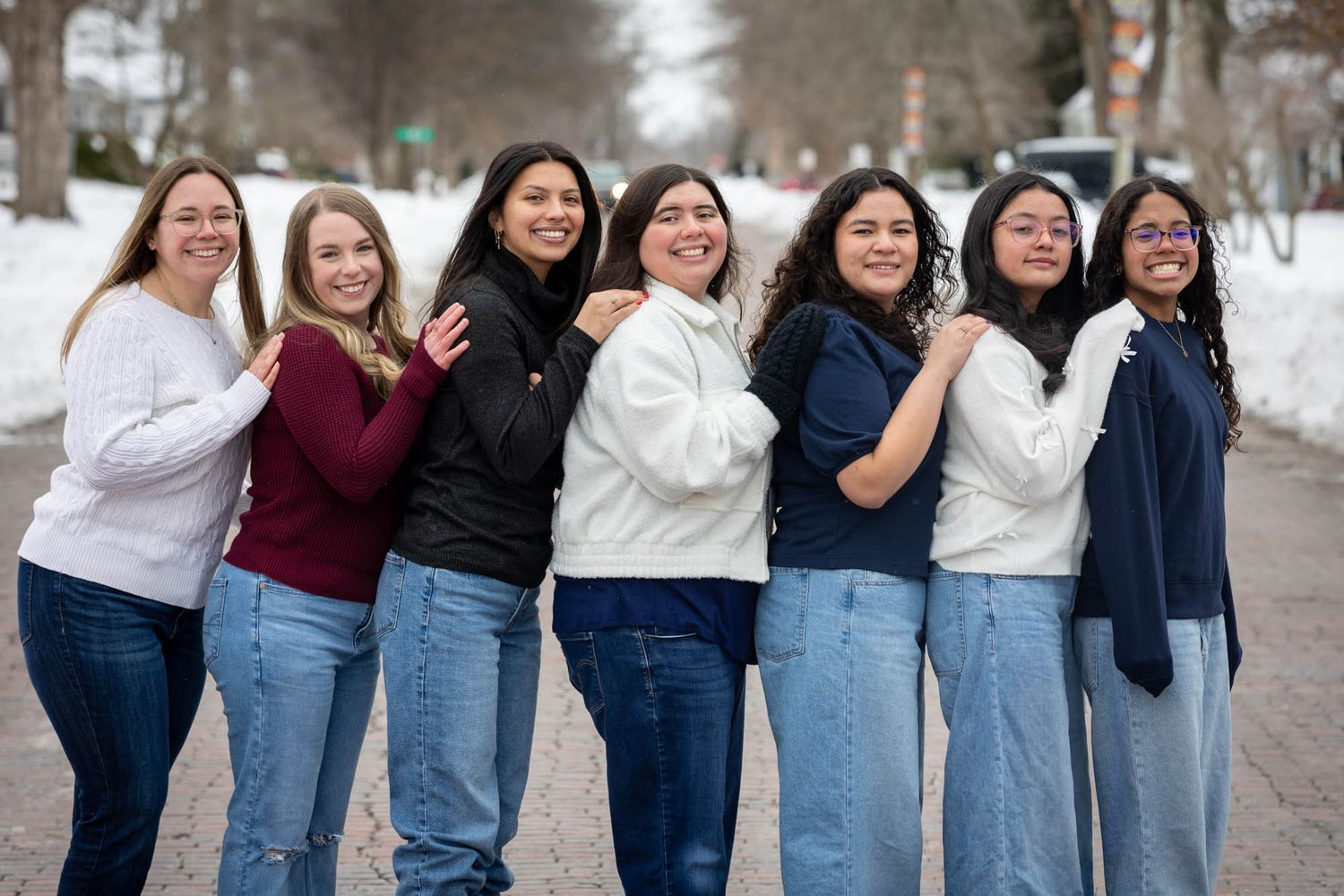 Group of seven smiling women posing together outdoors in winter with snow-covered ground in background.