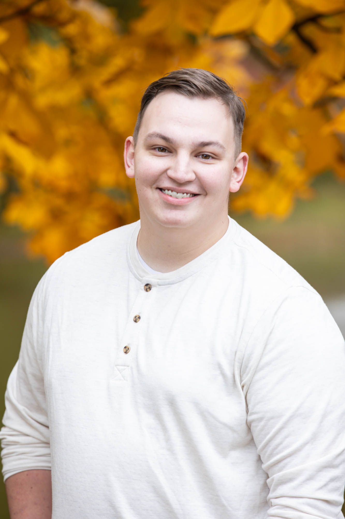 Smiling young man in a cream henley shirt posing outdoors with vibrant yellow autumn foliage behind him.
