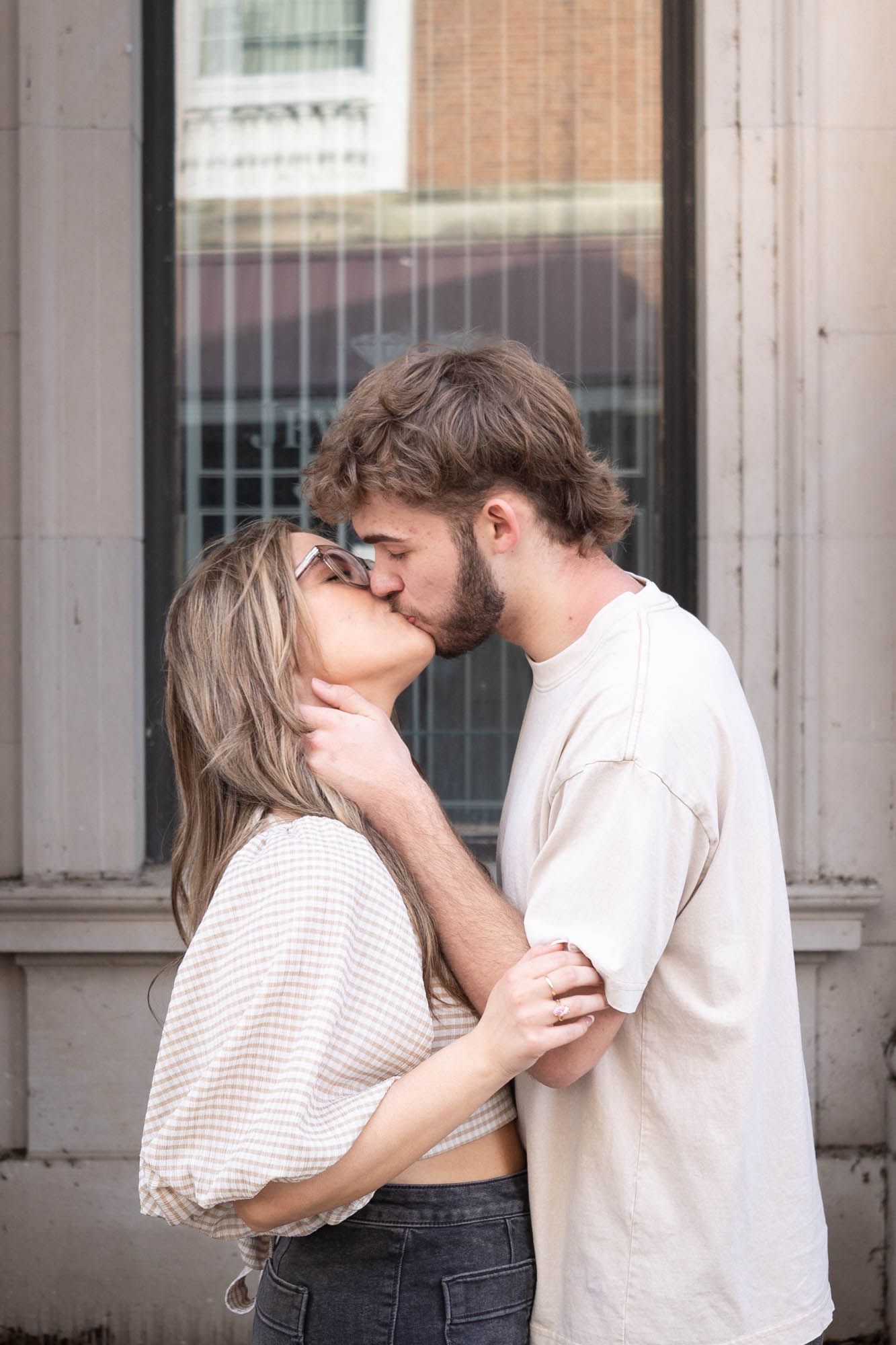 A couple shares a passionate kiss while embracing outdoors against a classic architectural building backdrop.