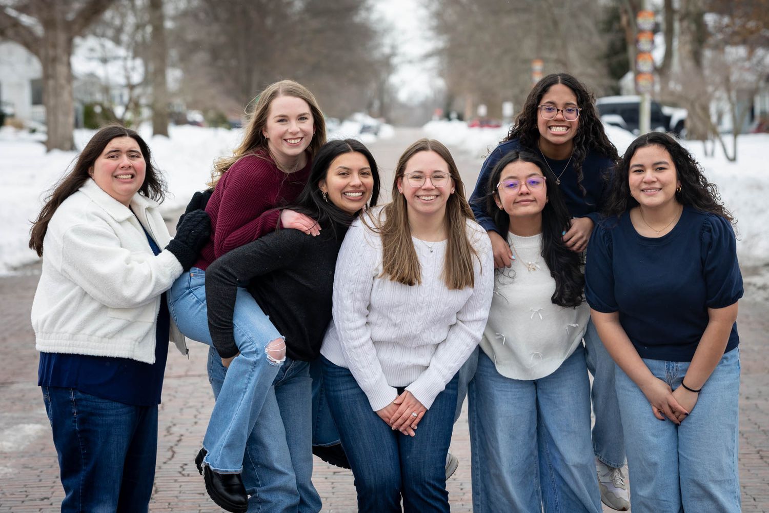 Group of seven smiling young women pose casually together on a snowy winter street.