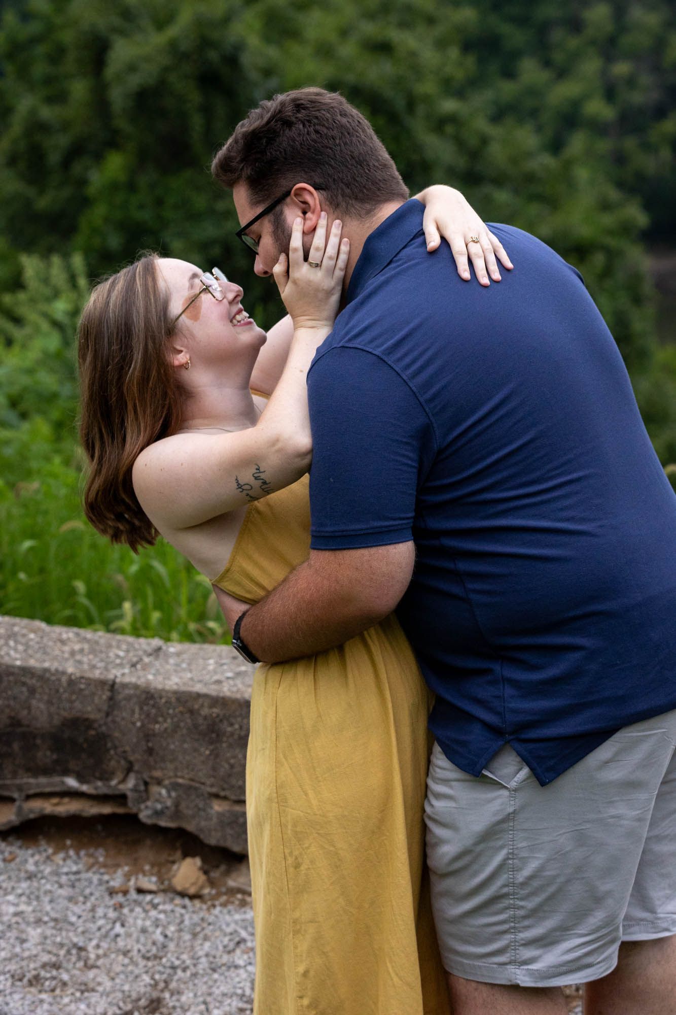 A couple shares a romantic embrace outdoors, woman in yellow dress and man in navy polo, smiling face to face.