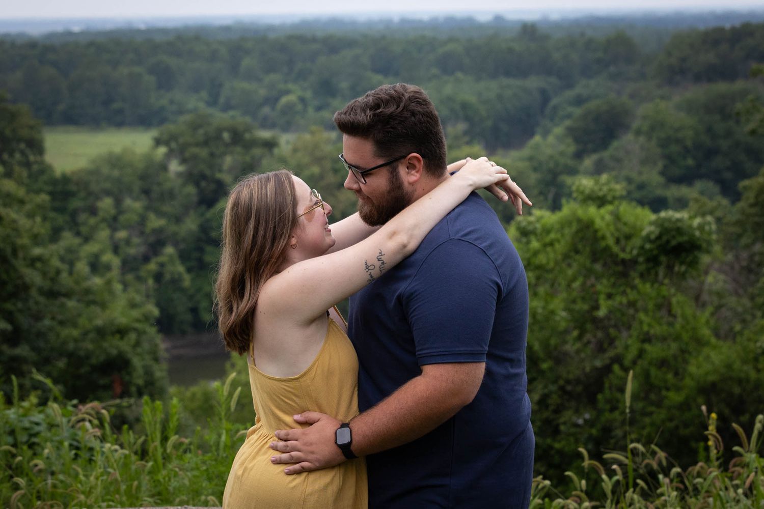 Couple embracing and smiling at each other outdoors with lush green forest landscape in the background.