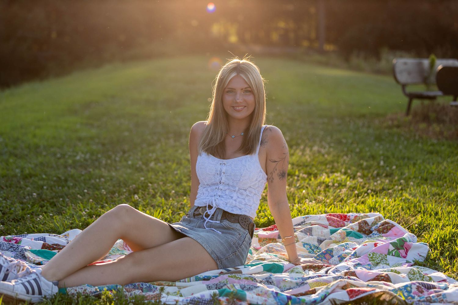 Young woman in white top and denim skirt sitting on a colorful quilt in a sunlit park.