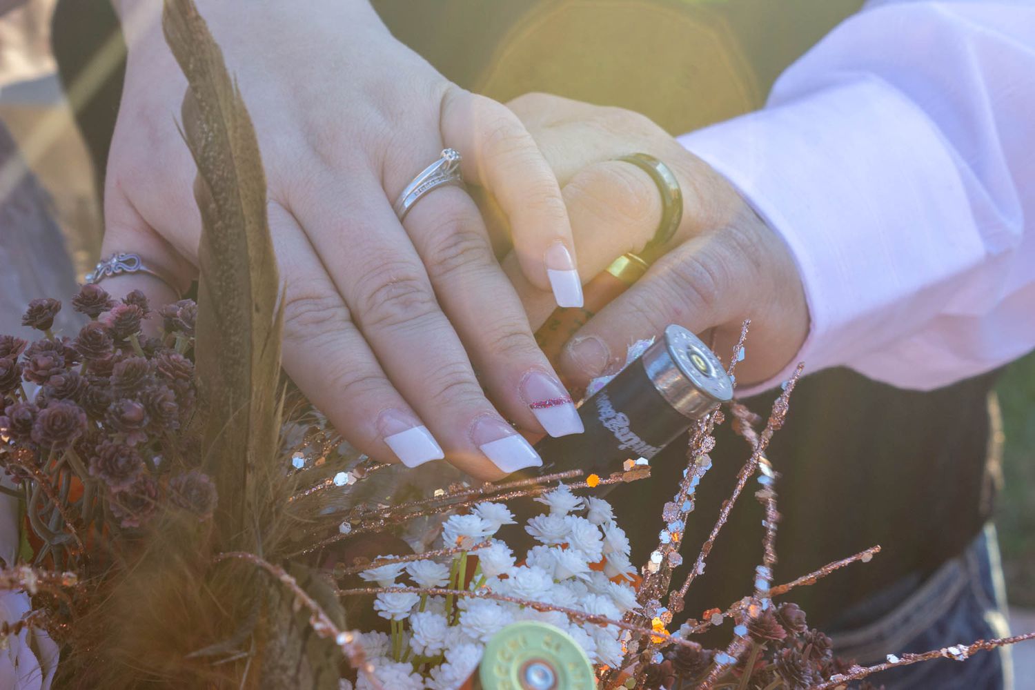 Close-up of bride and groom hands with wedding rings holding dried floral arrangement in sunlight.