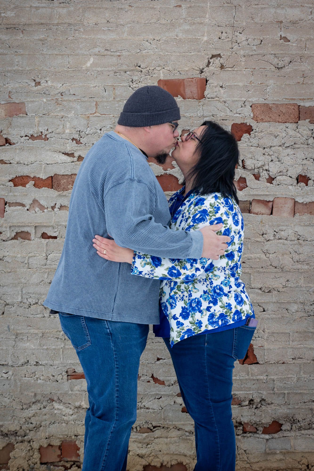A couple shares a romantic kiss while embracing in front of a weathered brick wall in winter.