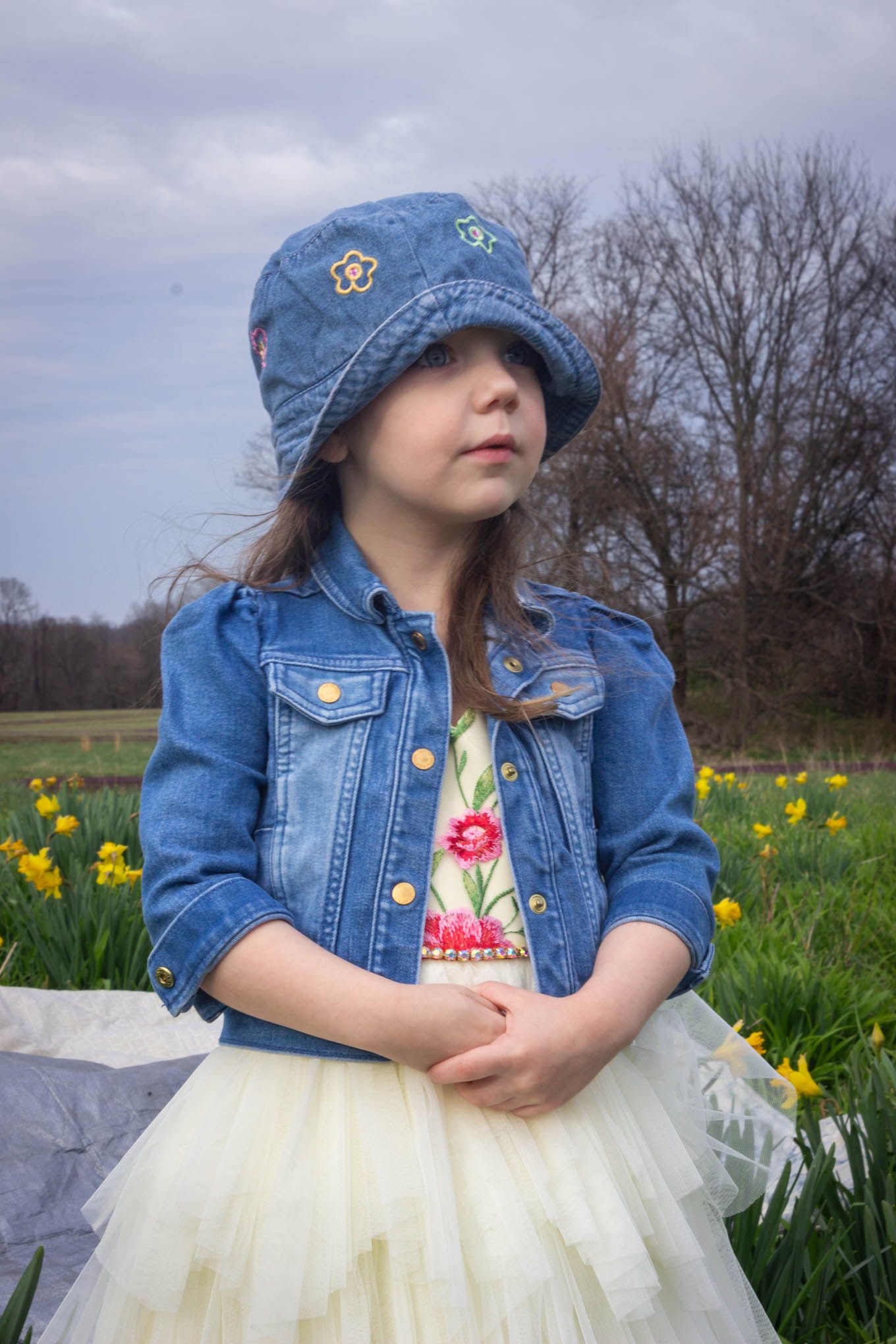 Young girl in denim jacket, bucket hat, and tulle skirt posing near yellow daffodils on cloudy day.