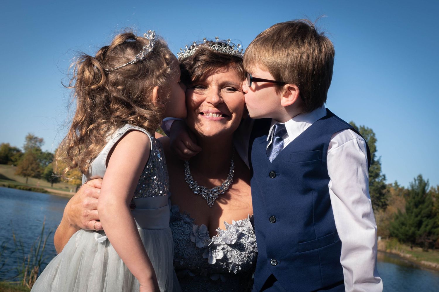Bride wearing tiara being kissed on cheeks by flower girl and ring bearer near a lake.