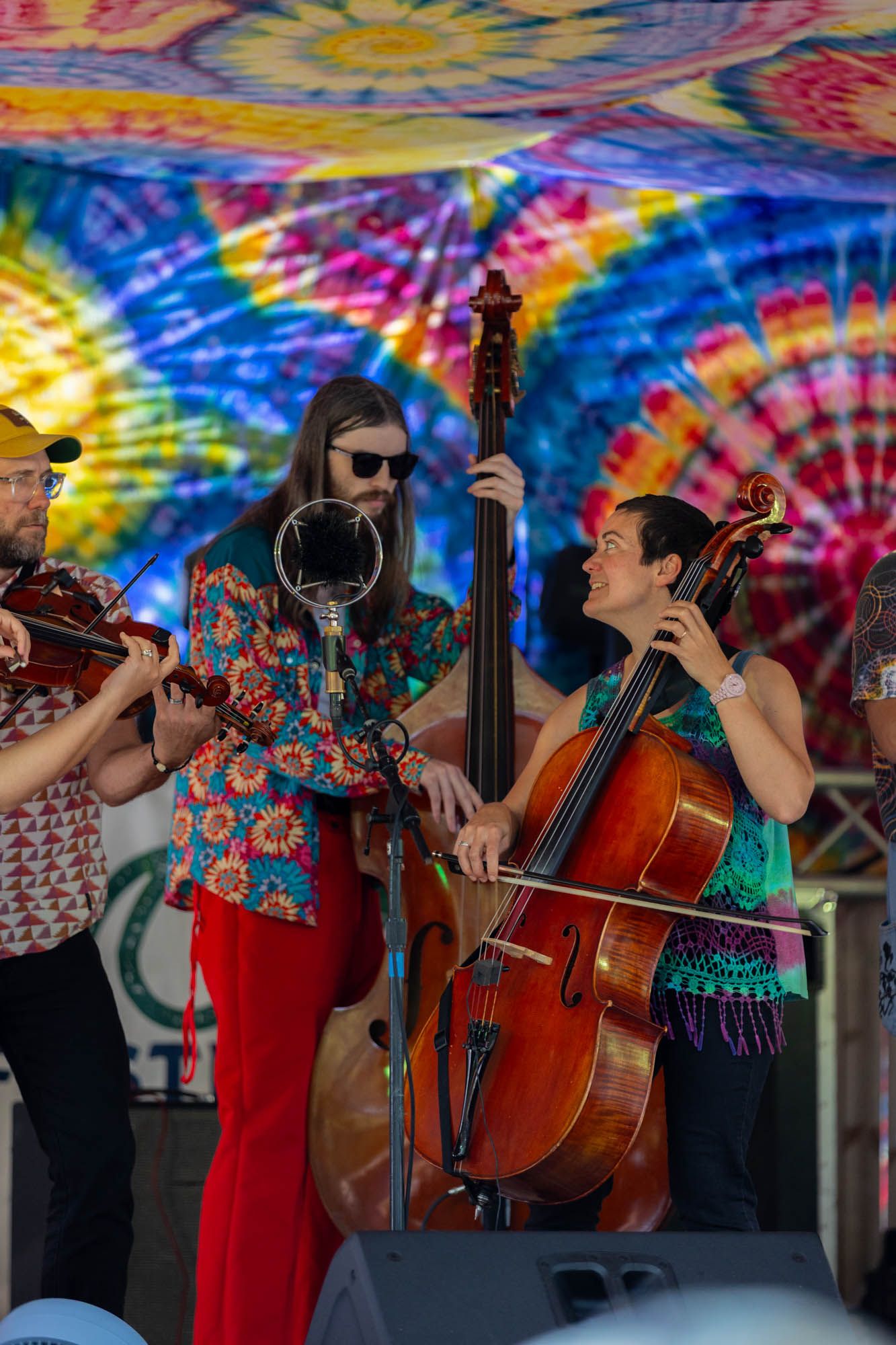 Musicians perform with double bass and cello on stage beneath a colorful tie-dye backdrop at an outdoor festival.