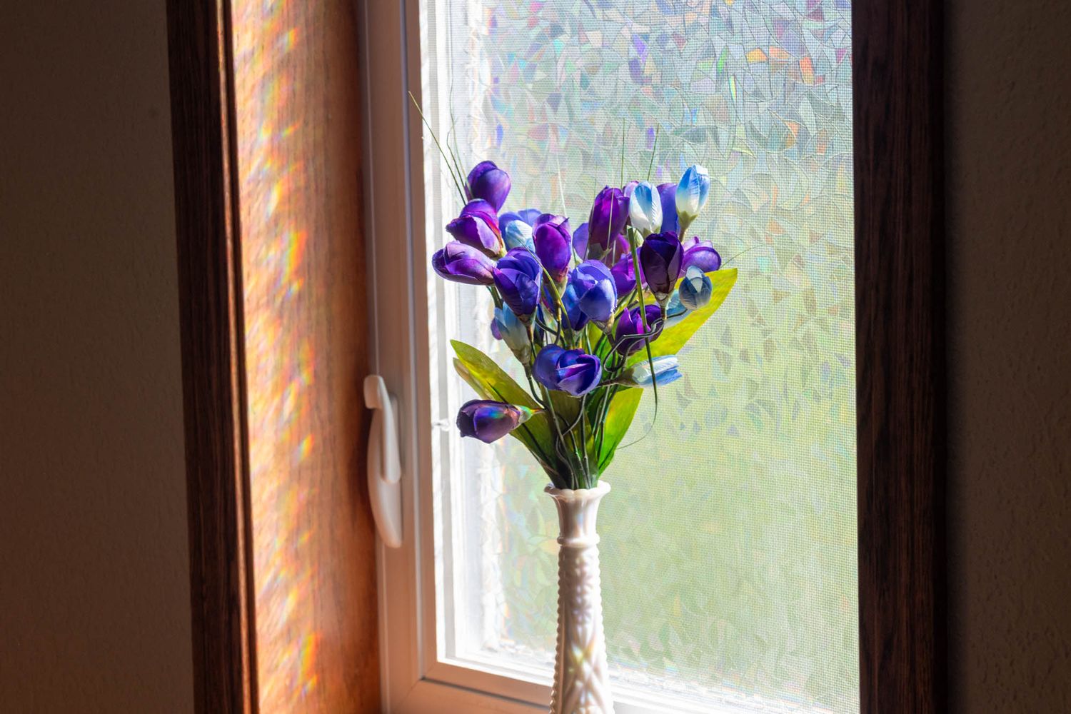 Purple tulip bouquet in white vase on windowsill with warm sunlight streaming through frosted glass.