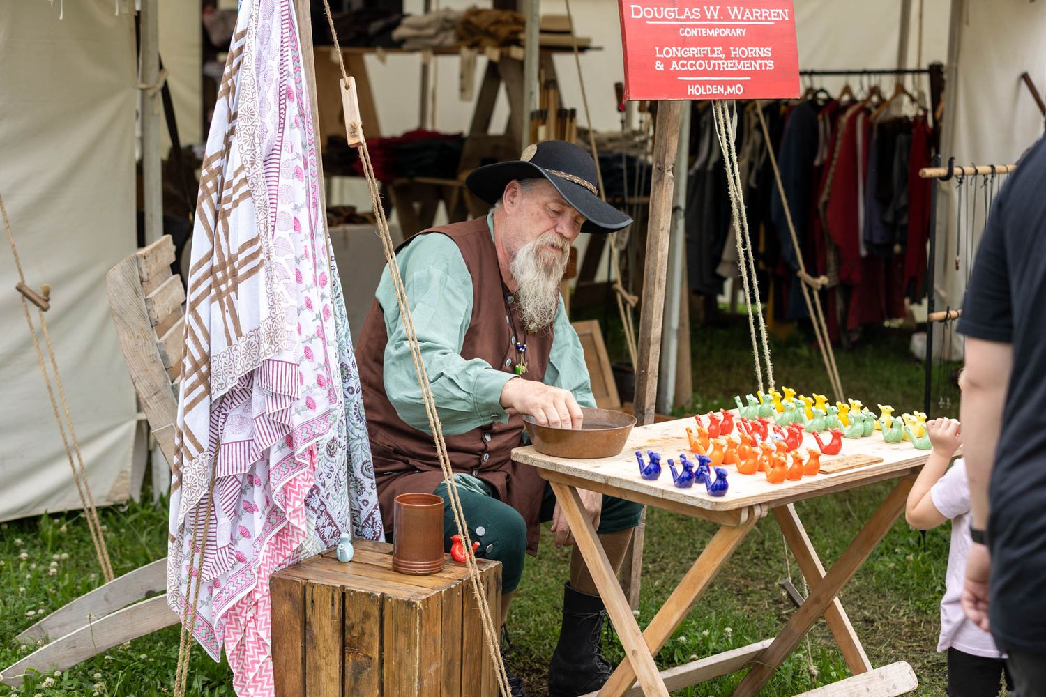 A bearded reenactor in period costume sells colorful glass chess pieces at an outdoor historical market tent.