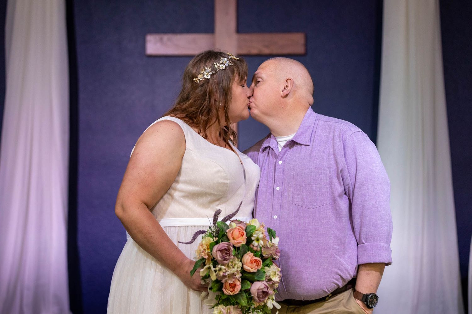 Bride and groom share first kiss at altar with wooden cross backdrop, bride holding colorful floral bouquet.