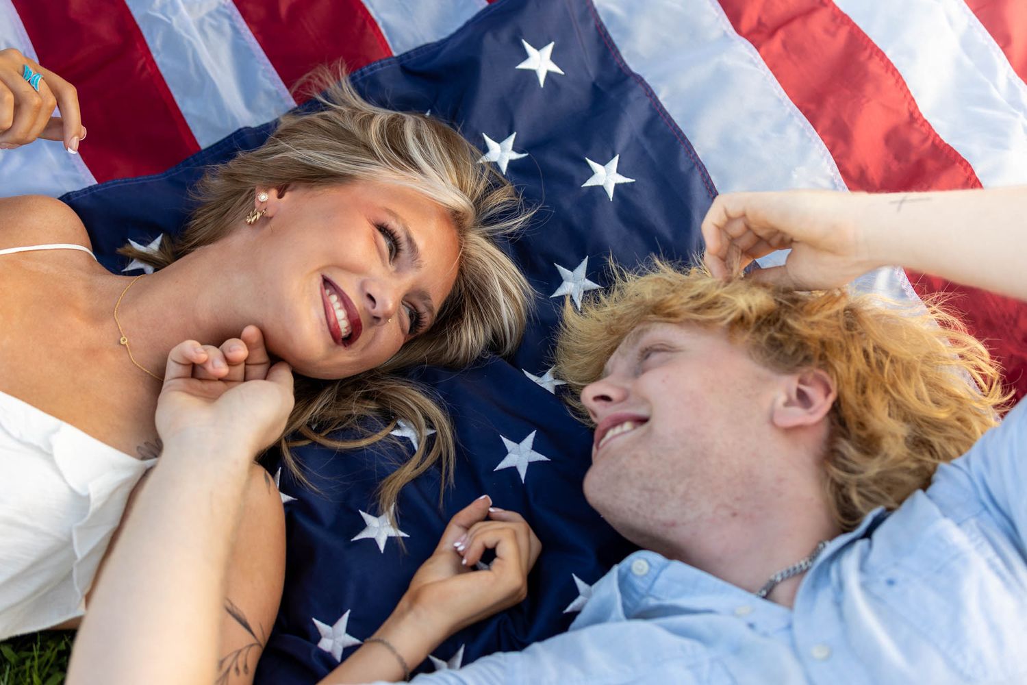 Two young people laughing and lying on an American flag outdoors on green grass.